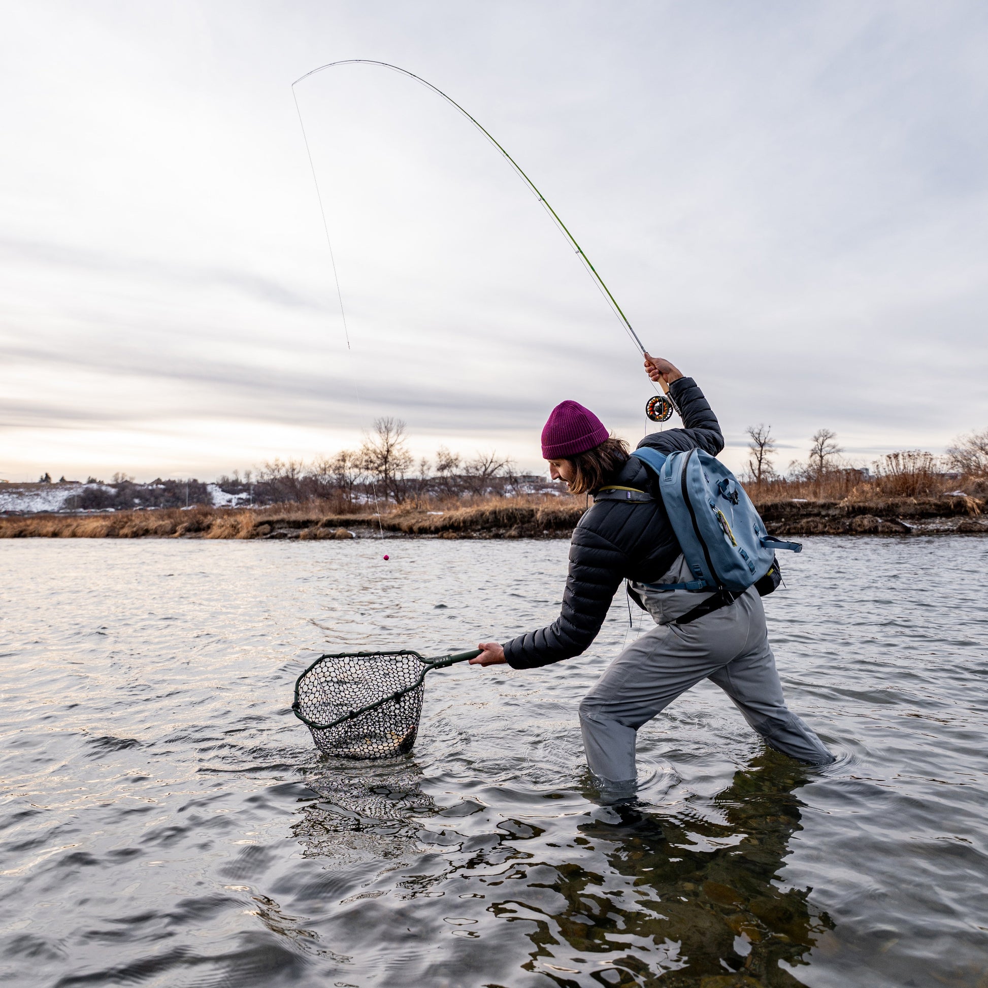 Person fly fishing in a body of water with a backpack and waders and landing net (global).