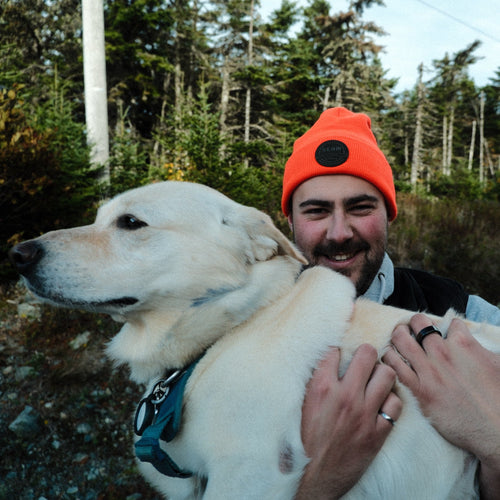 Man wearing an orange beanie with a seam logo, holding a white dog outdoors with trees in the background.