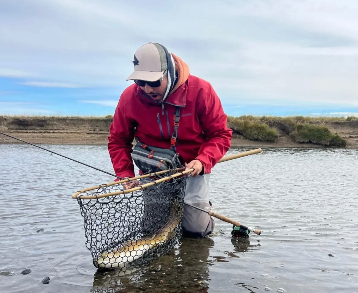 Person in a red jacket and gray waders holding a net in shallow water. Fly Fishing