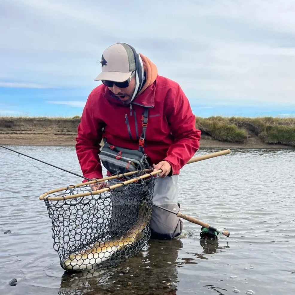 Person in a red jacket and gray waders fly fishing. They are holding a fishing net in shallow water (global).