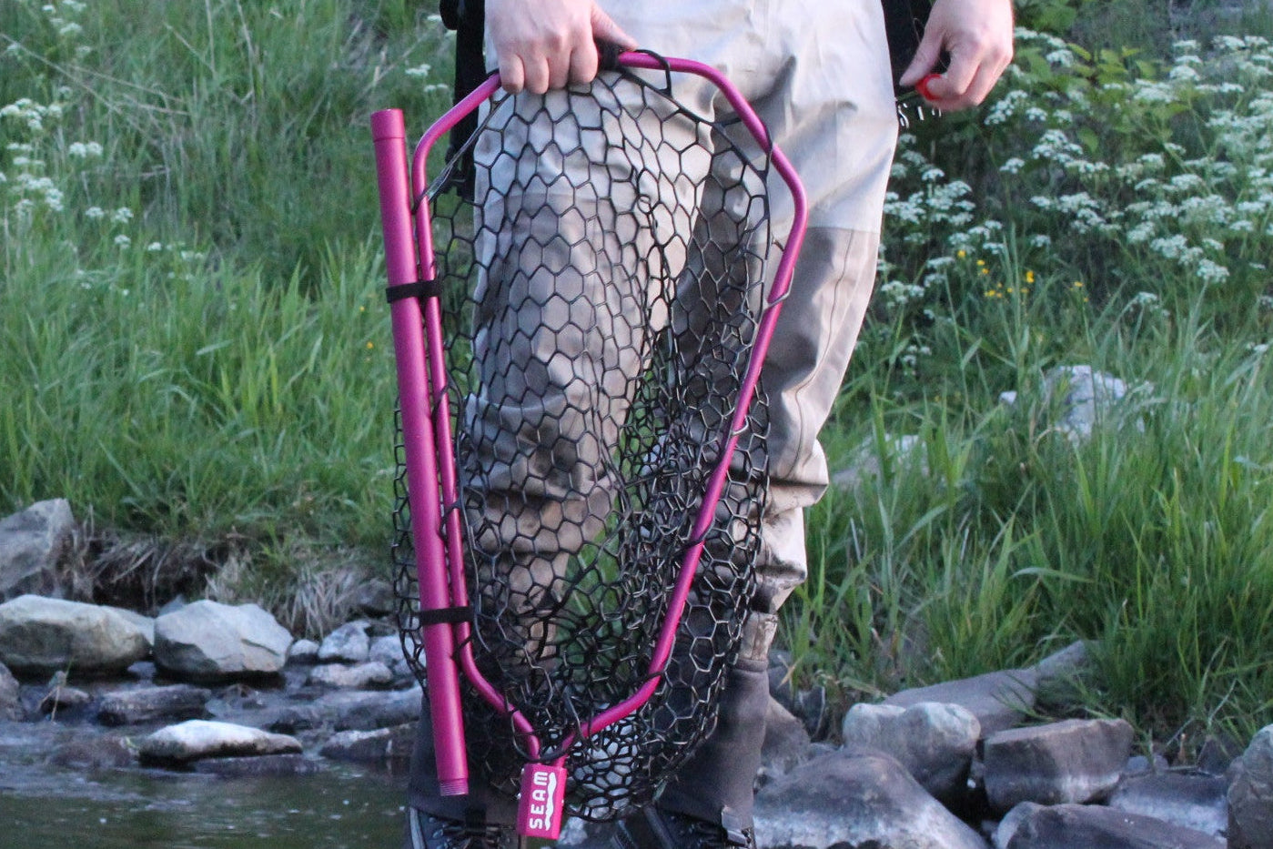 Person in waders holding a pink fishing net standing by a stream
