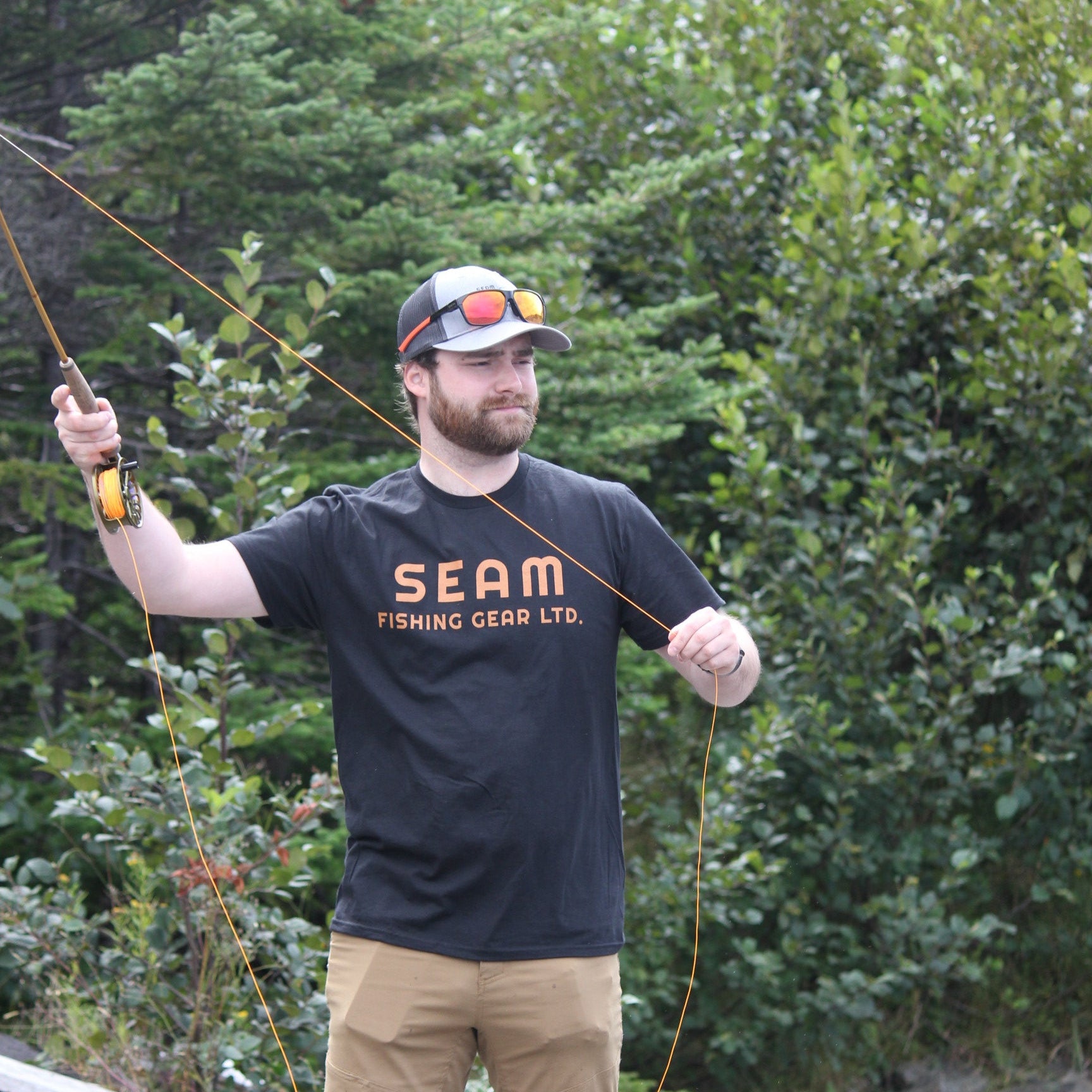 Man in a black shirt with 'SEAM' logo, tan pants, and a cap holding a fishing rod in a forest setting (global).