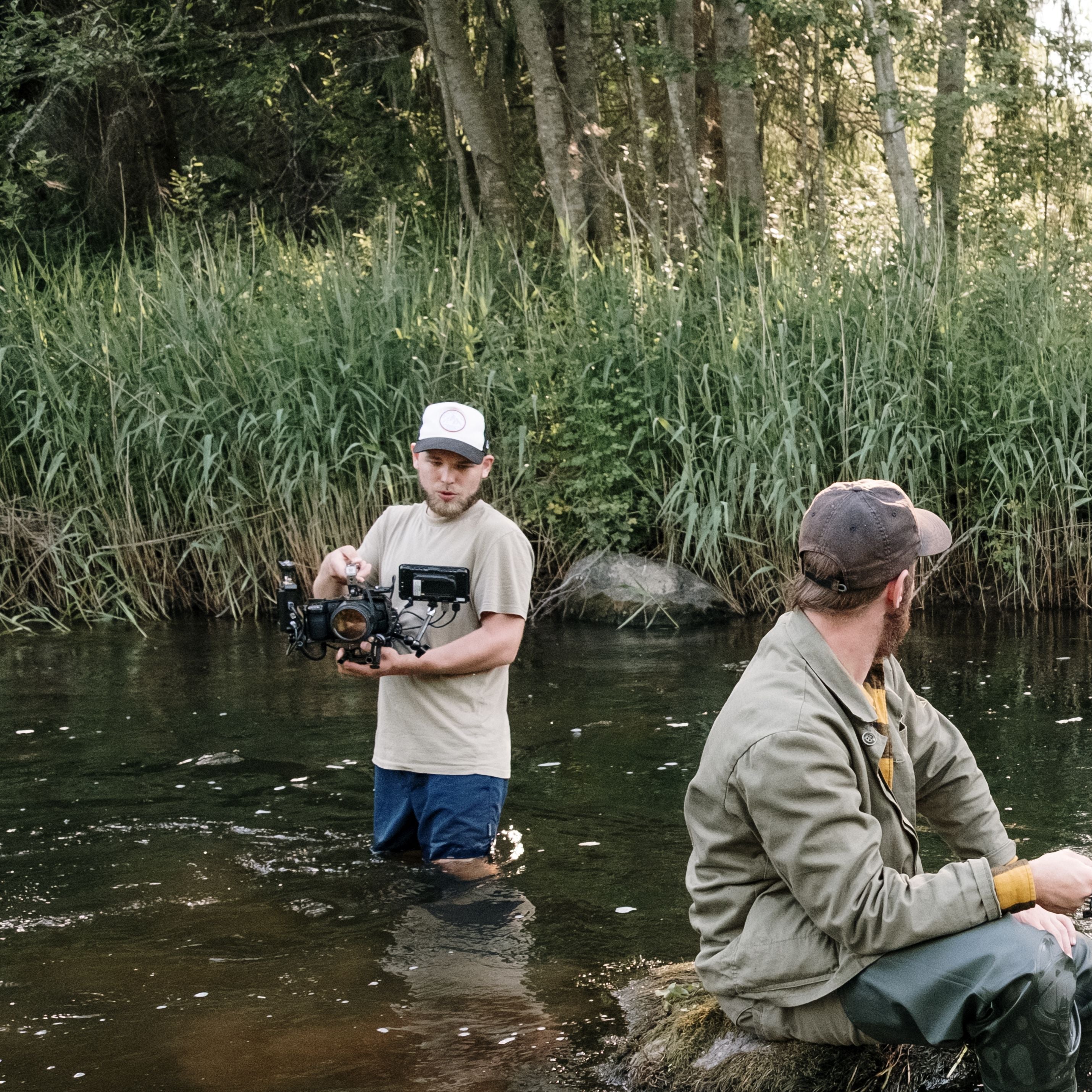 Two men fishing in a river with trees and reeds in the background